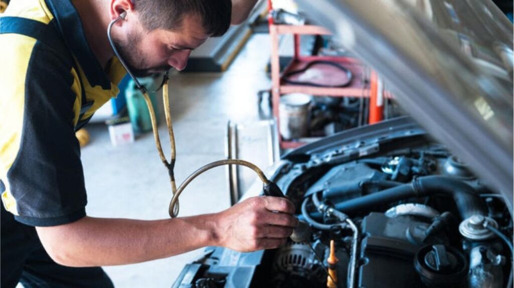 Mechanic holding a stethoscope to a car engine while the car runs in a dim workshop.