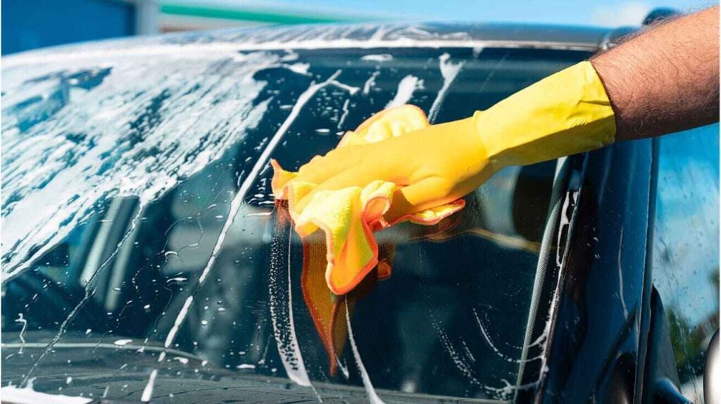 Alt Text: Person hand washing a car with a cracked windshield using a microfiber cloth and gentle water pressure.
